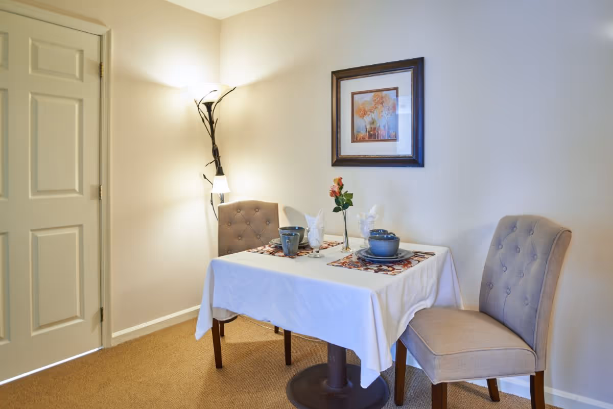 Small dining nook with a table set for two, two upholstered tufted chairs, a floor lamp, and framed artwork on a beige wall.