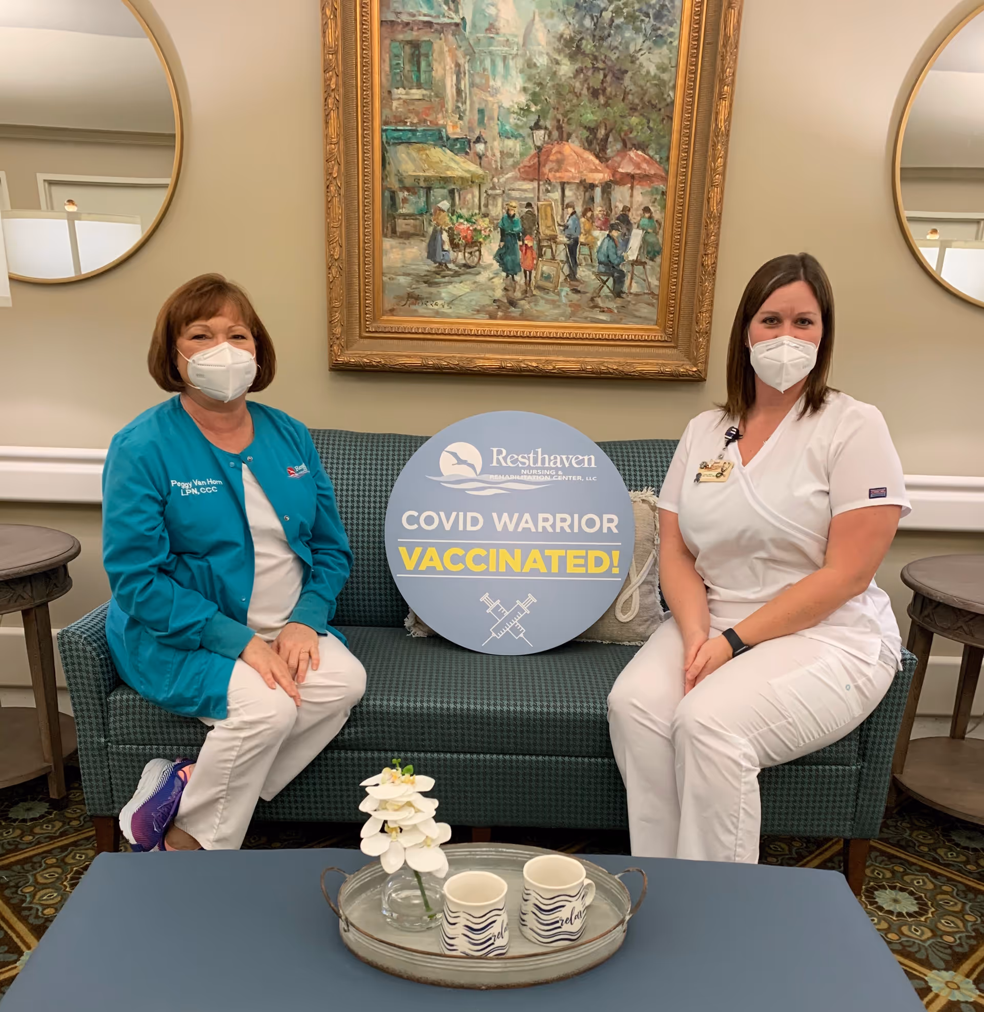 Two healthcare workers wearing masks sit on a green couch in a nursing facility lobby. They hold a round sign that reads 'Resthaven Nursing & Rehabilitation Center, LLC COVID WARRIOR VACCINATED!' Behind them is a framed painting of a street scene and two round mirrors on the wall. A coffee table with a tray holding two mugs and a small flower arrangement is in front of them.