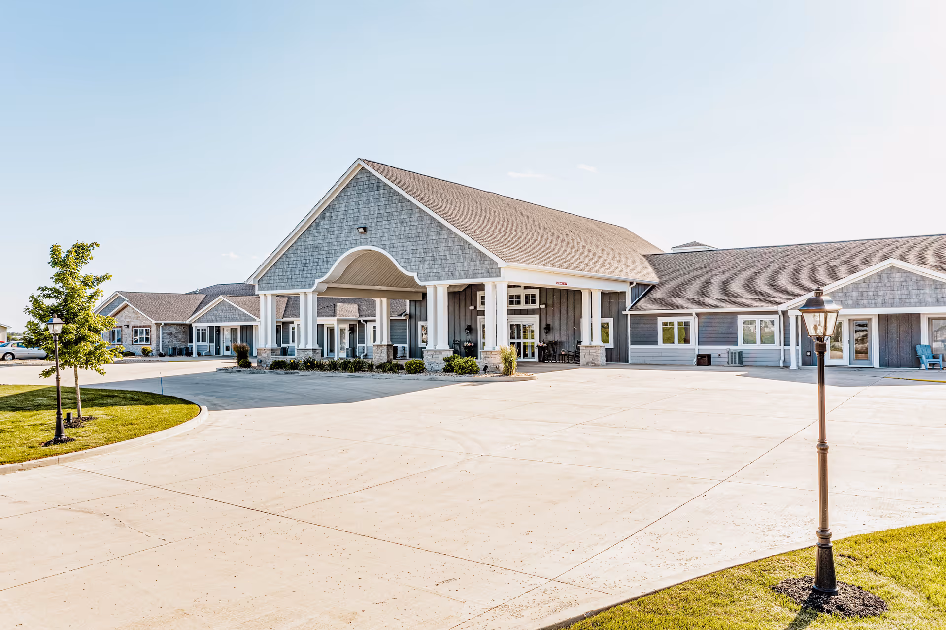 Exterior front view of Villas of Holly Brook Assisted Living & Memory Care facility in Jerseyville, IL, showing a large covered entrance with columns, gray siding, multiple windows, a paved driveway, and a lamp post on a grassy area.