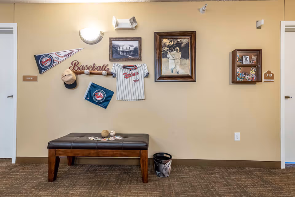 Interior hallway wall decorated with Minnesota Twins baseball memorabilia including pennants, a jersey, a baseball bat with the word 'Baseball', and a cap. There is a bench below the decorations with baseballs and cards on it. Two framed pictures hang on the wall, one showing a couple walking and the other a black and white photo of a house. There are two doors on either side of the wall labeled 'Laundry' and 'D10'.