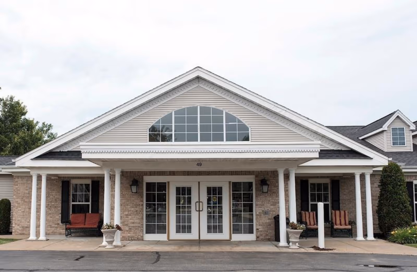 Front entrance of a senior living building with a gabled portico, white columns, double glass doors, and benches on either side.
