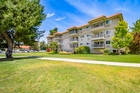 A multi-story residential building with balconies, surrounded by green grass, trees, and a sidewalk under a blue sky with some clouds.