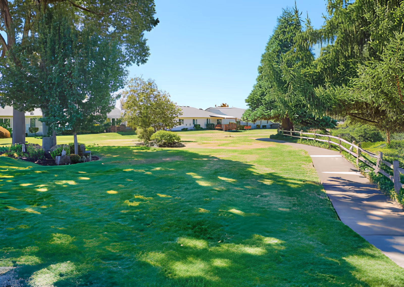 A sunny outdoor area at Elliott Residence featuring a well-maintained grassy lawn, several large trees providing shade, a paved walking path with a wooden fence on one side, and single-story buildings in the background under a clear blue sky.