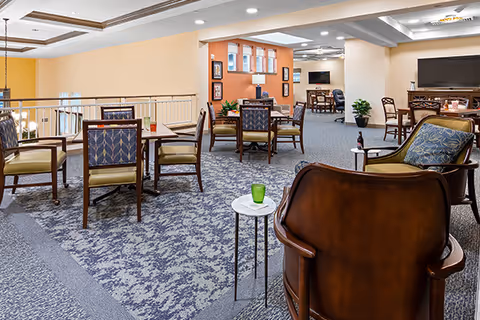 Spacious communal seating and dining area with multiple tables, chairs, and a TV in a senior living facility.