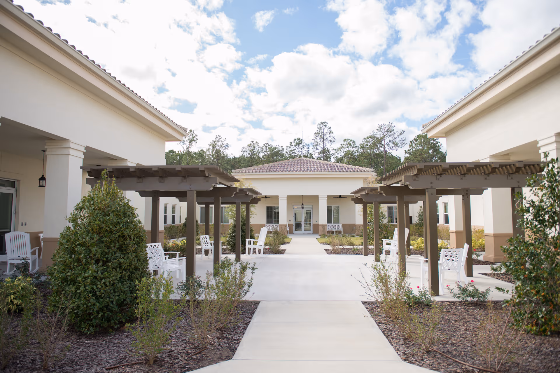 Outdoor courtyard area at Silver Creek facility with white chairs arranged under wooden pergolas, surrounded by bushes and plants, with a building entrance in the background under a partly cloudy sky.