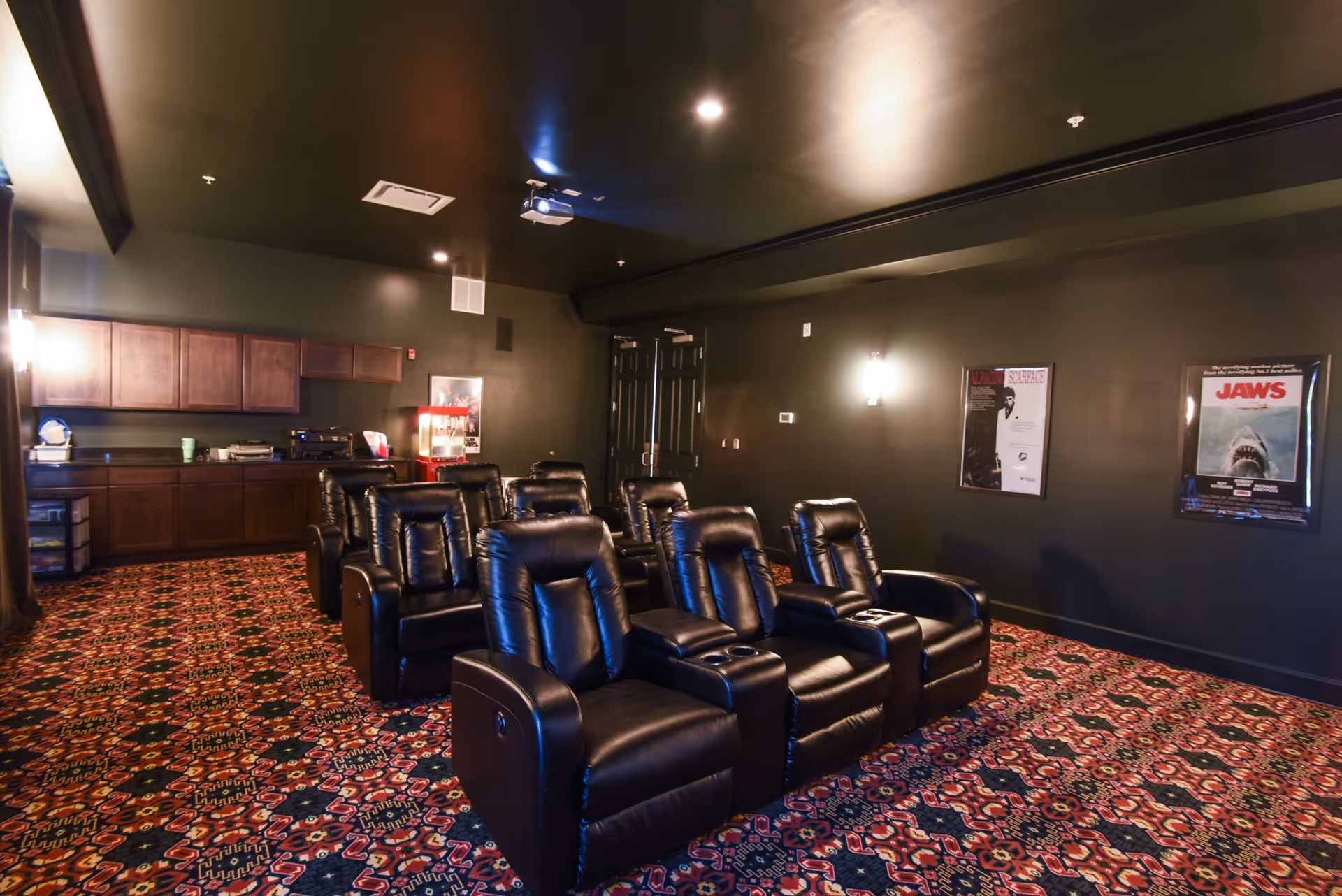 A small movie theater room with two rows of black leather recliner chairs facing a projector screen. The room has dark walls and ceiling, patterned carpet, and movie posters on the walls. There is a popcorn machine and cabinetry along the back wall.