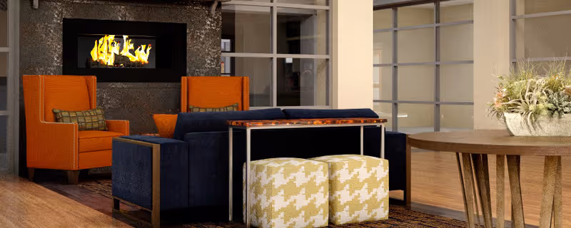A cozy living room area featuring two orange armchairs, a dark blue sofa, two patterned ottomans, a wooden table with metal legs, a modern fireplace with visible flames, and a large window with multiple panes. There is also a decorative plant on the wooden table.