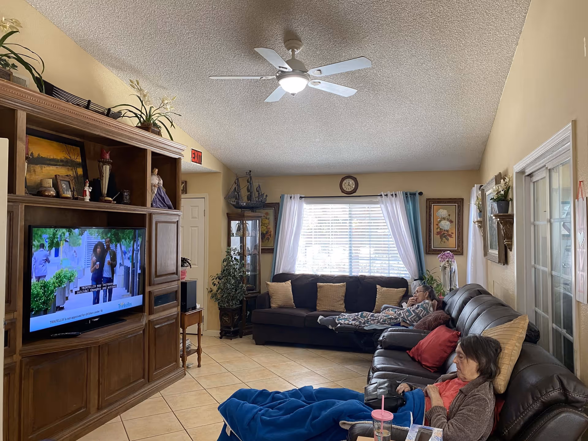 Living room with a large wooden entertainment center and TV, sofas with throw pillows, a ceiling fan, and two seated residents.