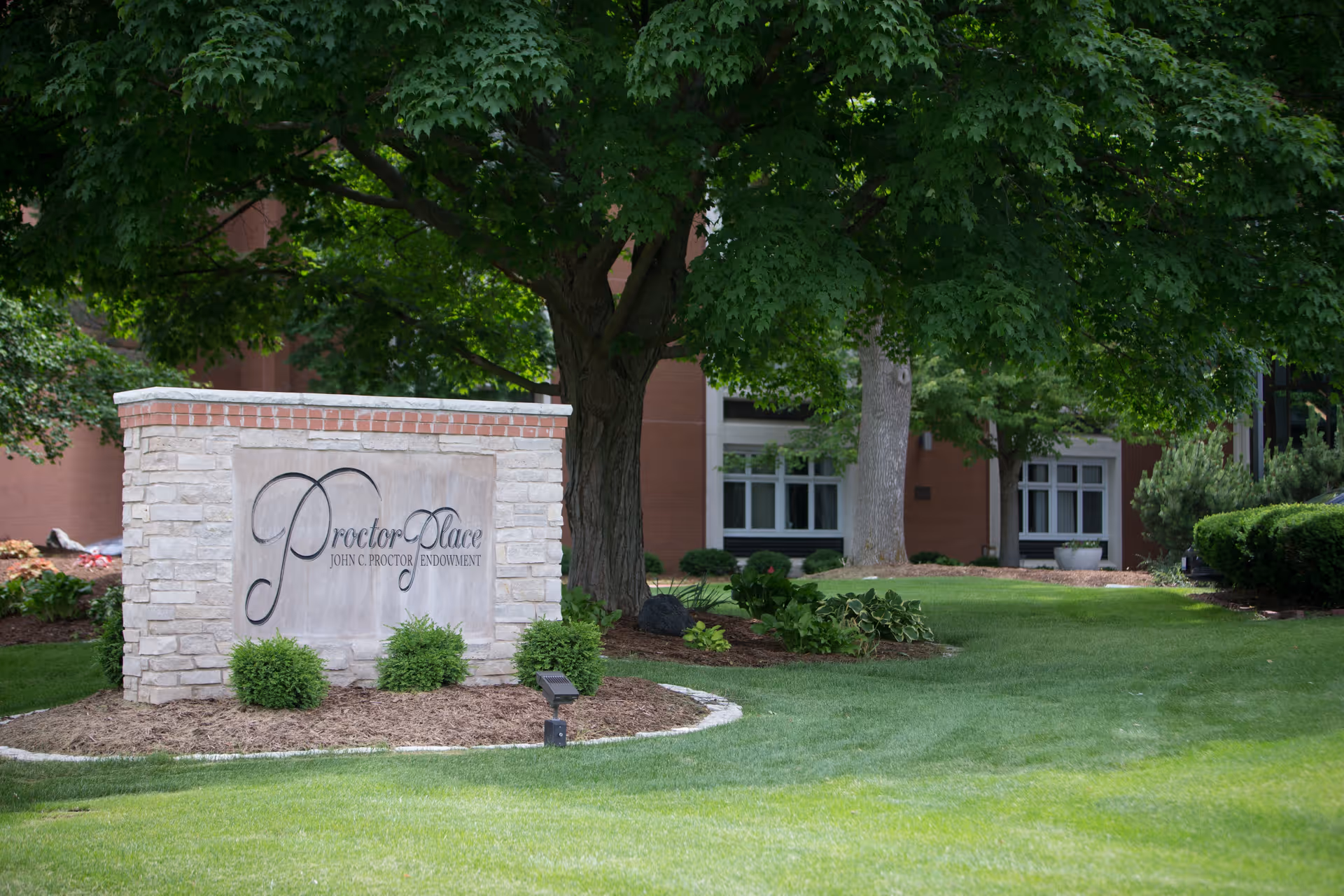 Stone sign for Proctor Place John C. Proctor Endowment surrounded by green grass, shrubs, and trees with a building partially visible in the background.