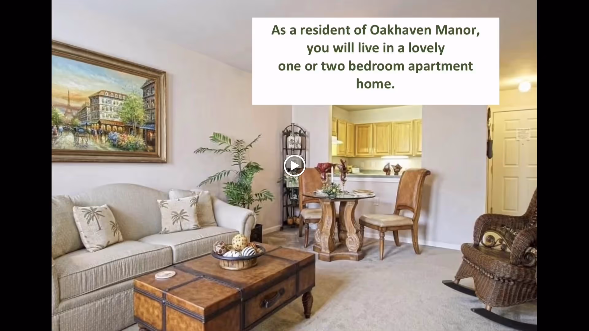 A cozy living room and dining area in an apartment at Oakhaven Manor. The living room features a beige sofa with palm tree patterned pillows, a wooden coffee table with decorative items, a large framed painting on the wall, and a potted plant. The dining area has a round wooden table with four chairs and is adjacent to a kitchen with wooden cabinets. A wicker rocking chair is also visible near the entrance door.
