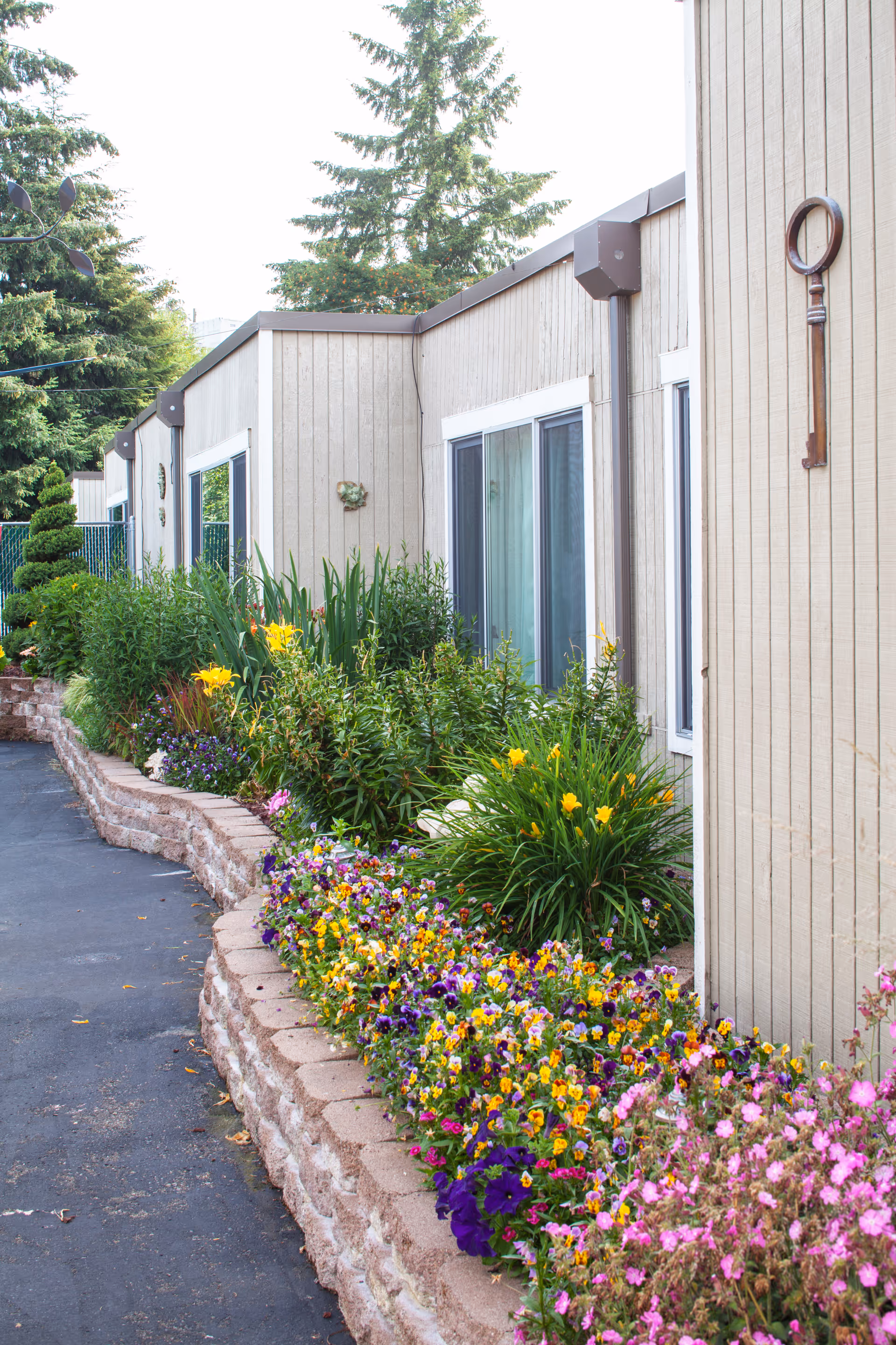 A landscaped garden area outside a beige building with large windows. The garden features a variety of colorful flowers and green plants bordered by a low stone wall. A large decorative key is mounted on the building's exterior wall.
