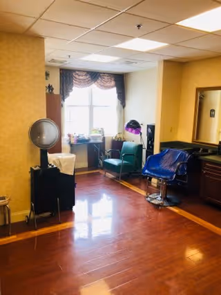Interior of a hair salon area with a salon chair covered in a blue cape, a green chair, a large mirror on the wall, a window with decorative curtains, and various hair styling equipment.