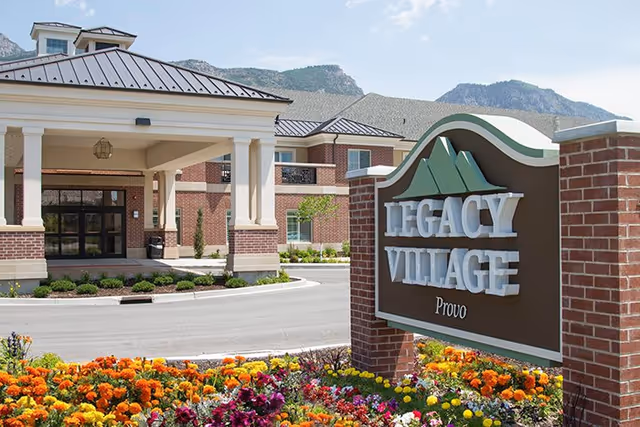 Exterior view of Legacy Village of Provo senior living facility with a large entrance canopy, brick building, and a landscaped flower bed in front of the facility sign. Mountains are visible in the background under a clear sky.