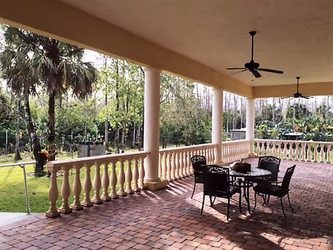 Covered outdoor patio with columns, ceiling fans, brick pavers, and a table and chairs overlooking a grassy yard and trees.