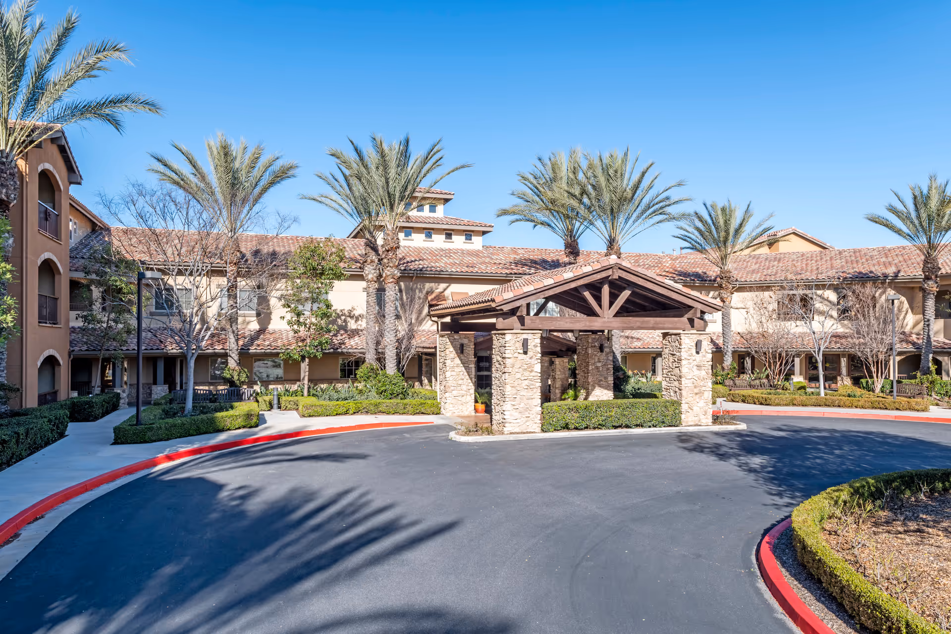 Exterior view of Valencia Terrace senior living facility showing a circular driveway with a covered entrance supported by stone pillars, surrounded by palm trees and landscaped greenery under a clear blue sky.