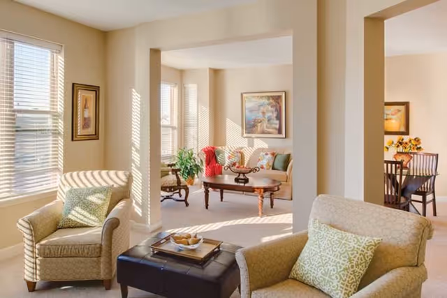 A bright and cozy living room area with two beige armchairs featuring patterned green cushions, a black ottoman with a tray on top, and a view into an adjacent sitting area with a beige sofa, colorful cushions, a wooden coffee table, a plant, and framed artwork on the walls. Sunlight streams through the windows with white blinds.