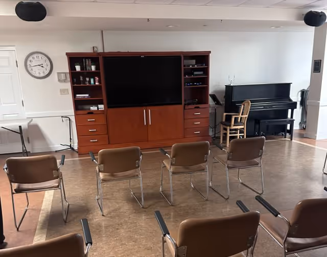 A room with a large wooden entertainment center holding a flat-screen TV, several shelves with items, and multiple drawers. In front of the entertainment center are two rows of brown chairs with metal legs facing the TV. To the right, there is a black upright piano with a wooden chair in front of it. A clock is mounted on the wall to the left, and the floor is a mix of brown and beige tiles.