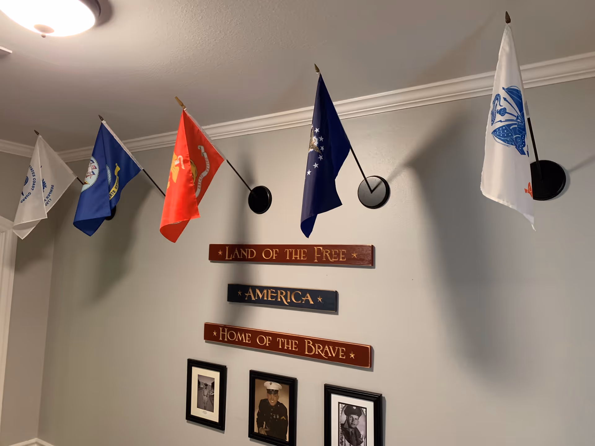Interior wall display featuring five military flags mounted on angled black holders, three wooden plaques with the inscriptions 'LAND OF THE FREE', 'AMERICA', and 'HOME OF THE BRAVE', and three framed black and white photographs of military personnel beneath the plaques.