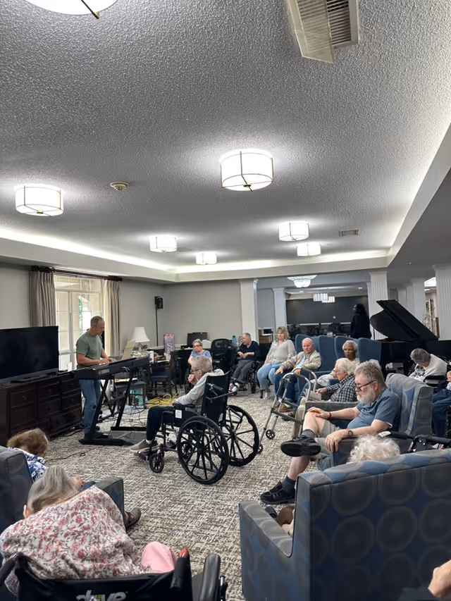 A group of elderly residents seated in a common room of an assisted living facility, some in wheelchairs and others in armchairs, attentively listening to a man playing an electronic keyboard. The room is well-lit with ceiling lights and has a carpeted floor, a large window with curtains, a television, and a grand piano in the background.