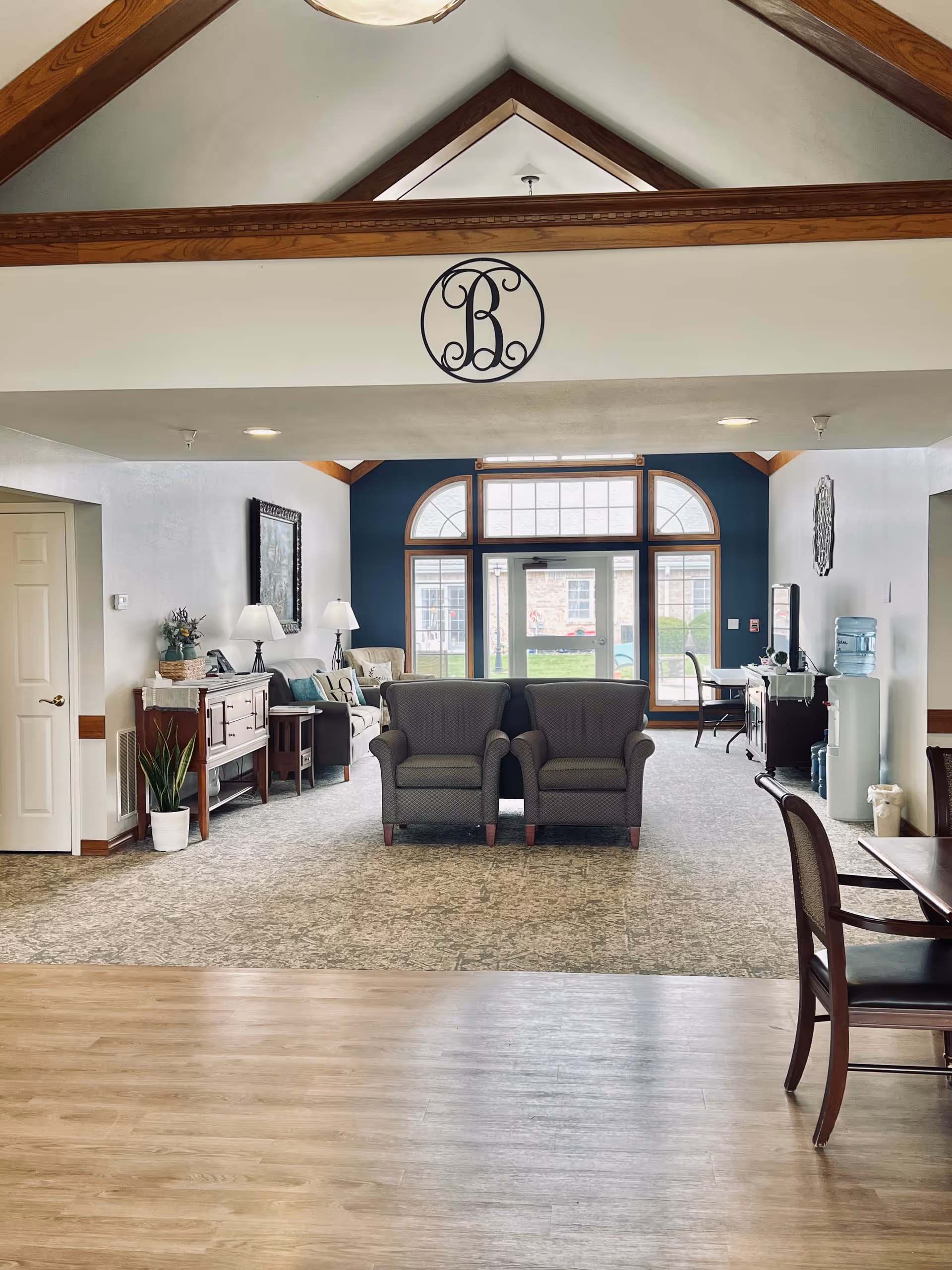Bright communal sitting area with two armchairs facing large arched windows, vaulted ceiling with wooden beams, side tables, lamps and a small dining table.