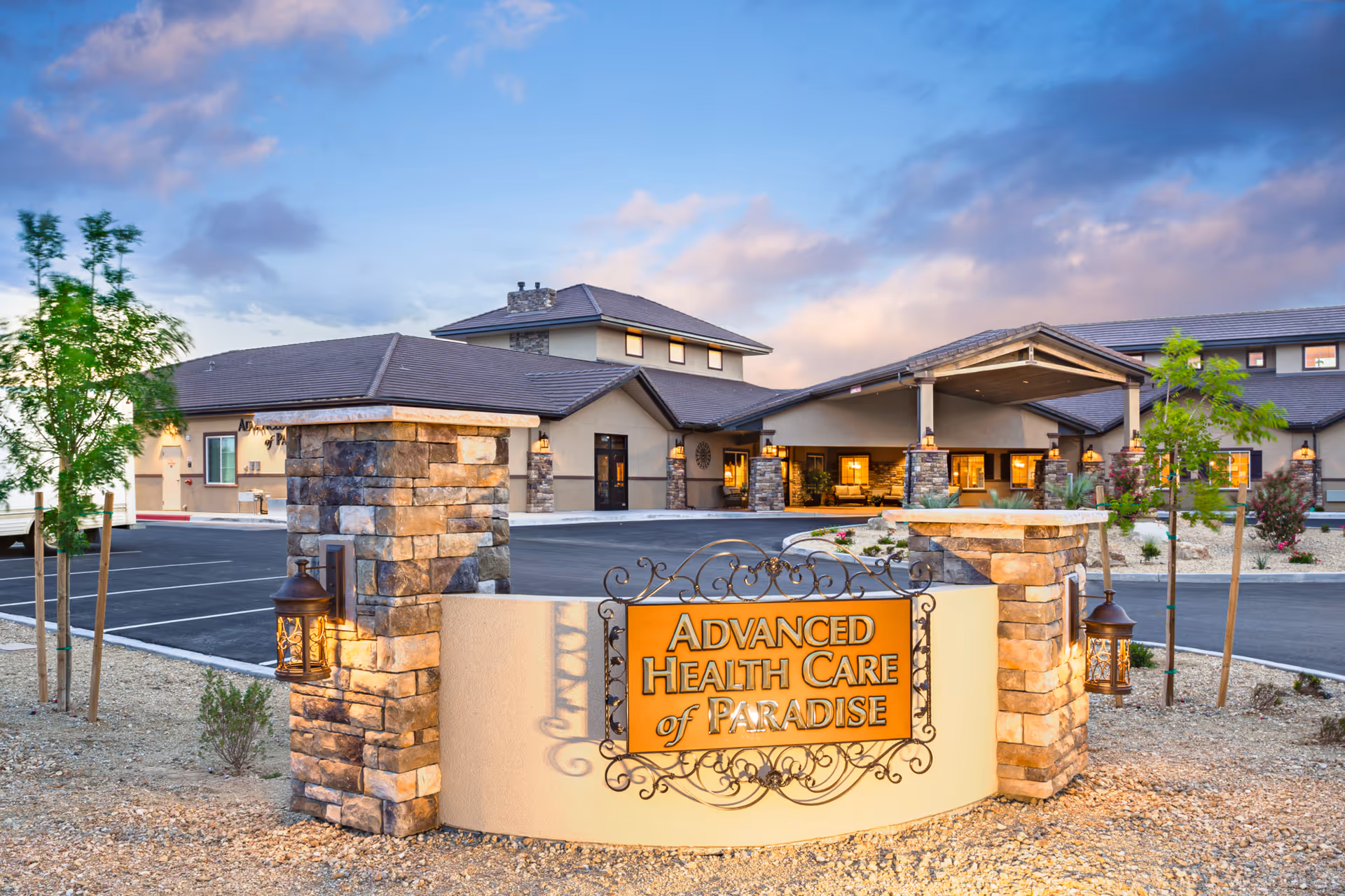 Exterior view of Advanced Health Care of Paradise facility at dusk, showing a stone and stucco sign with the facility name in front of a large building with multiple windows and a covered entrance, surrounded by a parking lot and landscaped with small trees and shrubs.