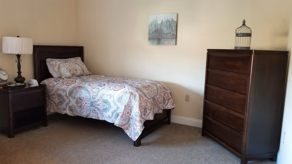 Furnished bedroom with a twin bed covered in a patterned quilt, a nightstand and lamp to the left, and a tall wooden dresser to the right.