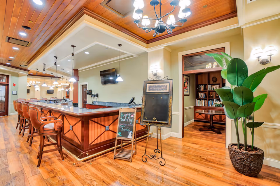 Interior view of a senior living facility's dining area featuring a wooden bar with high chairs, a chandelier, and pendant lights. There is a chalkboard sign with holiday greetings and a large potted plant near an entrance to a room with bookshelves and a table.