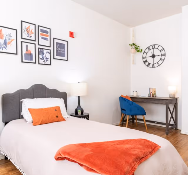 Bright furnished bedroom with a single bed, gray headboard, orange throw and pillow, bedside lamp, gallery wall, and a desk with a blue chair and wall clock.