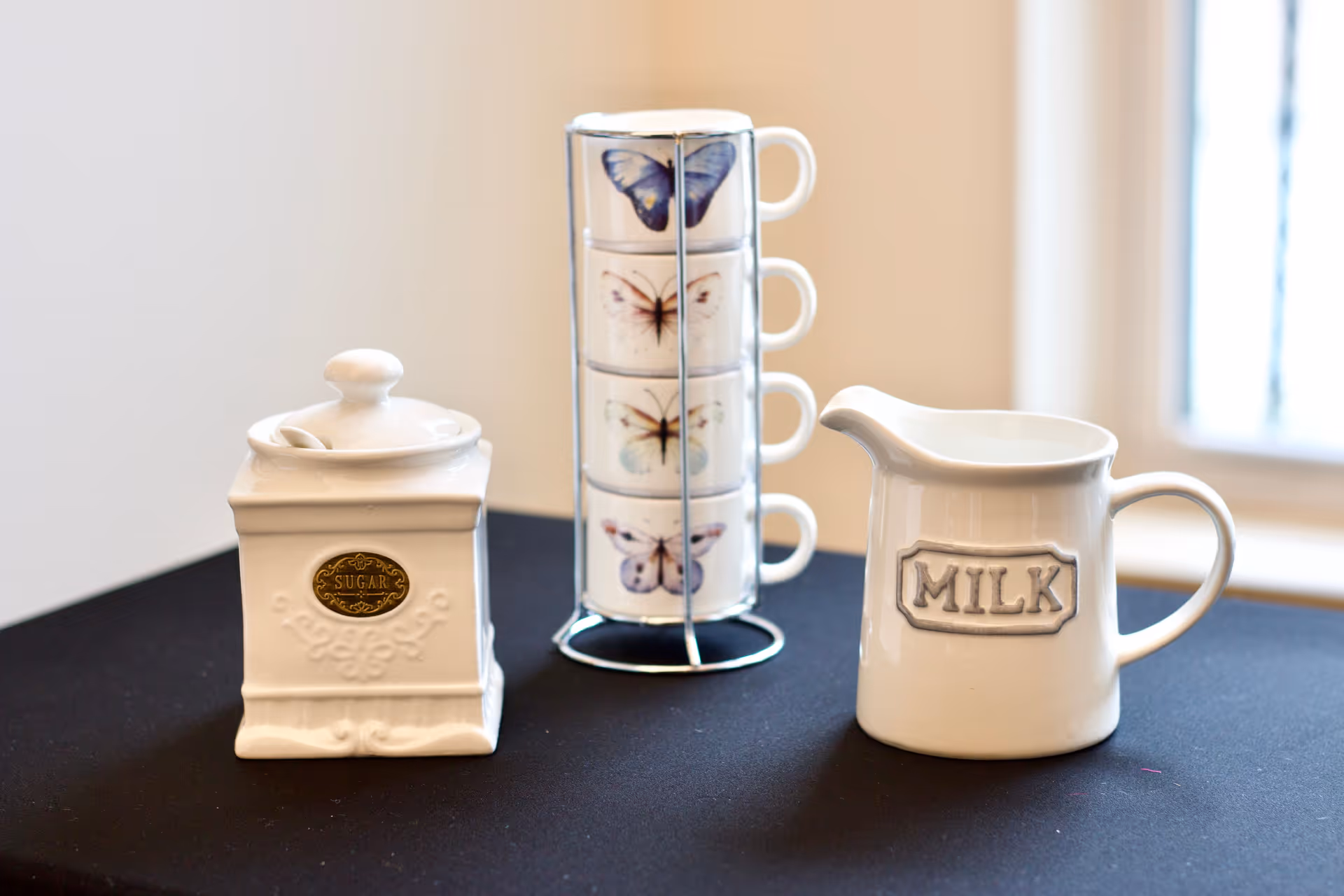 White sugar jar, a stack of butterfly-decorated teacups, and a white milk pitcher labeled "MILK" arranged on a black tablecloth.