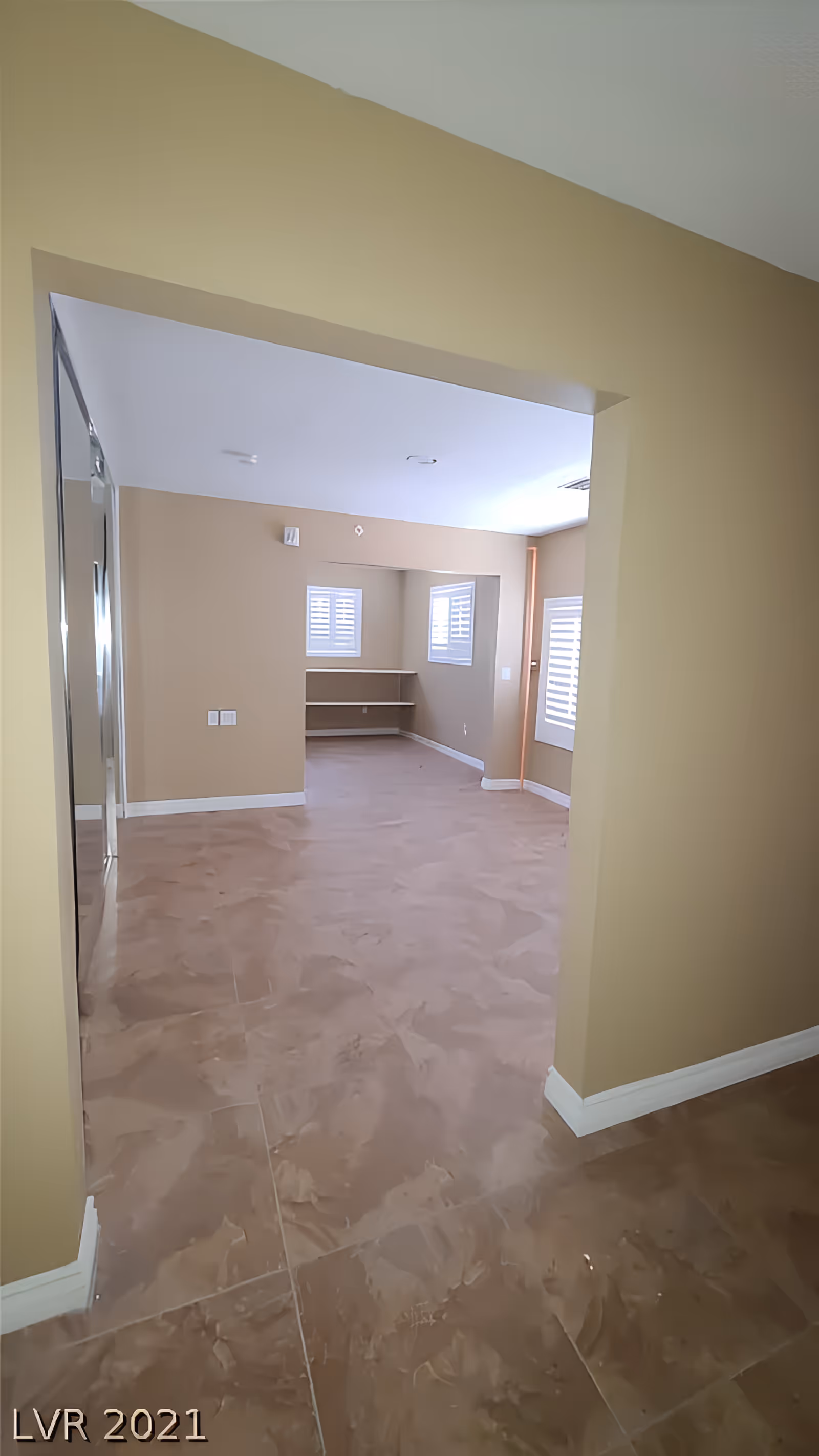 Empty interior room with beige walls and brown marbled floor tiles. The room has two small windows with white shutters and built-in corner shelves. The ceiling is white with recessed lighting fixtures.
