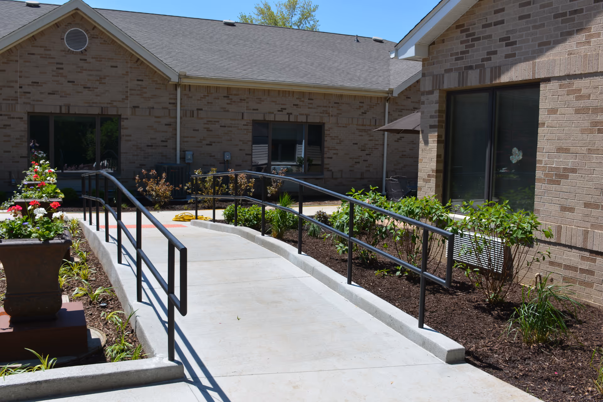 Outdoor view of a paved walkway with black metal handrails leading to the entrance of a brick building. There are flower beds with green plants and blooming flowers on either side of the walkway, and a large planter with red and white flowers on the left side. The building has multiple windows and a sloped roof under a clear blue sky.