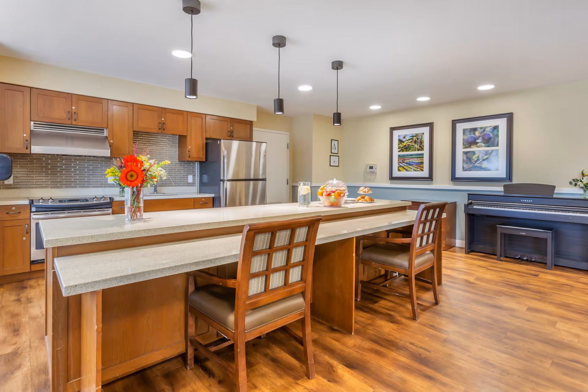 A modern kitchen area with wooden cabinets, stainless steel refrigerator and stove, and a long island countertop with two wooden chairs. The countertop has a vase with flowers, a glass of water with lemon, a bowl of fruit, and a plate of pastries. The room has wooden flooring, pendant lights hanging from the ceiling, and two framed pictures on the wall. A black Yamaha piano with a bench is positioned against the far wall.