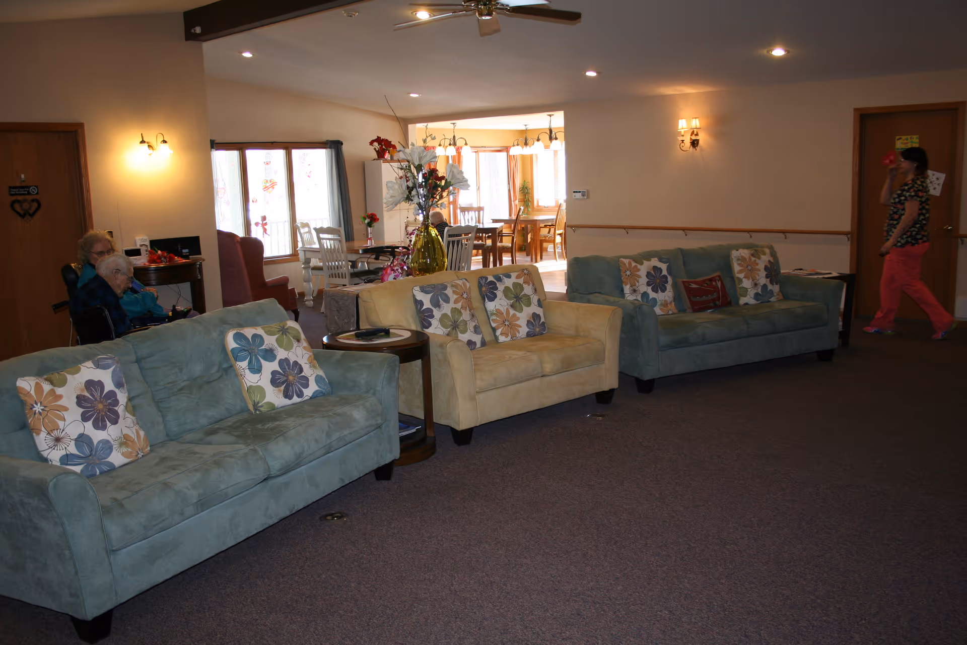 Communal lounge with several upholstered sofas and floral pillows facing an open dining area, with a vase of flowers on a side table and people in the background.