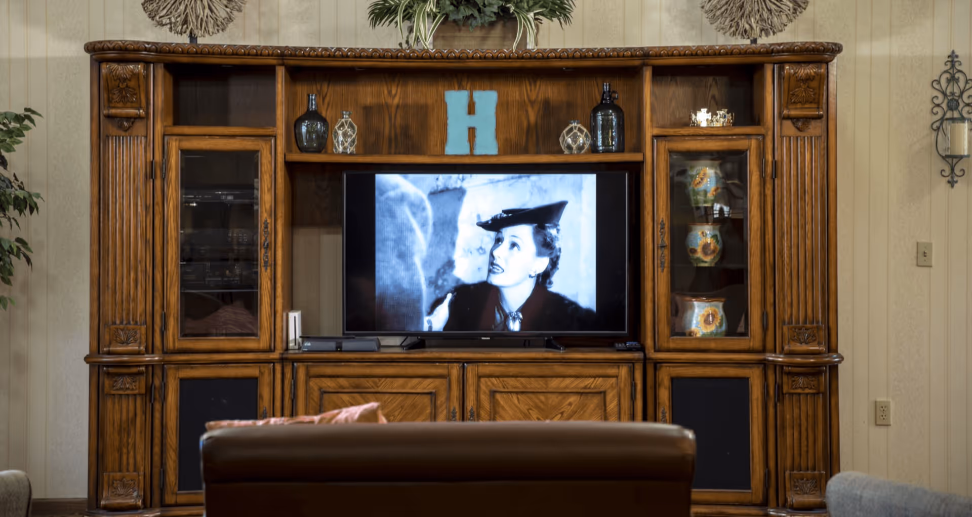 A wooden entertainment center with decorative carvings holds a flat-screen TV displaying a black-and-white movie scene featuring a woman in a hat. The unit has glass cabinets with decorative vases and bottles, and a large blue letter H on the top shelf. In front of the entertainment center is a brown leather chair and part of a gray chair is visible on the right. The room has beige wallpaper and a wall-mounted candle holder on the right.