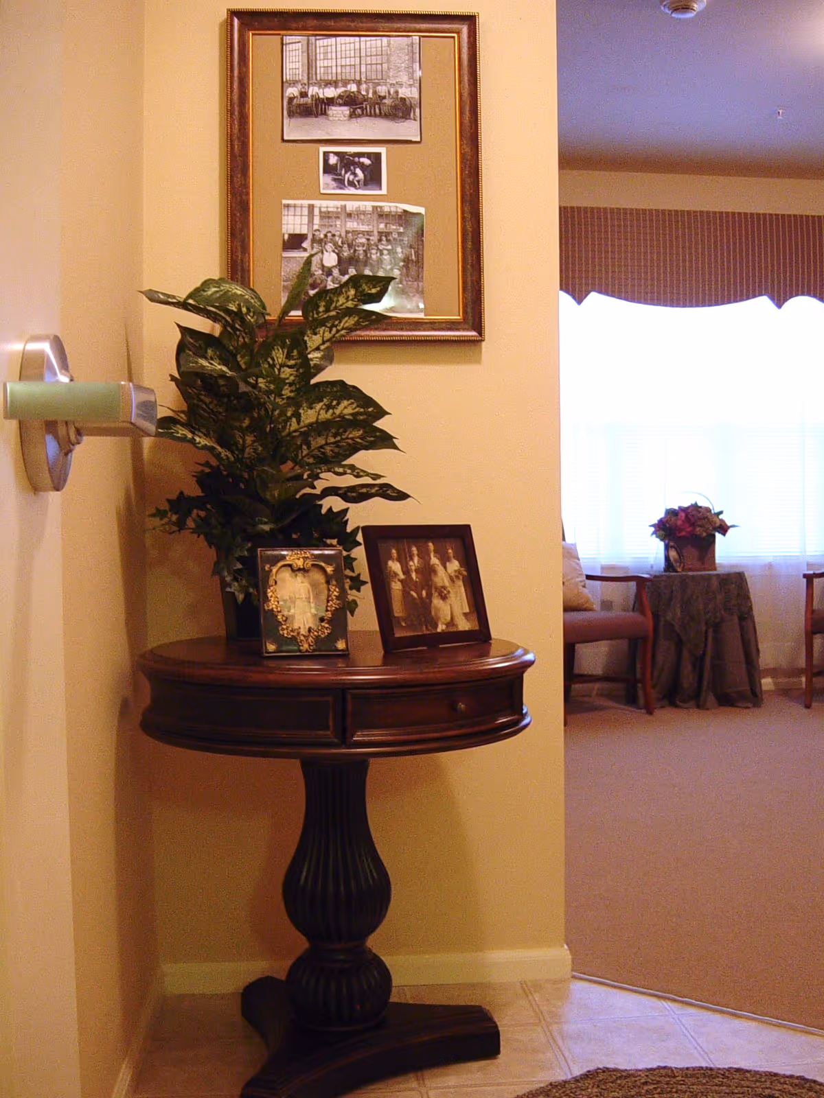Round wooden side table topped with a potted plant and framed photos against a wall, opening to a sunlit sitting area with chairs and a small table.