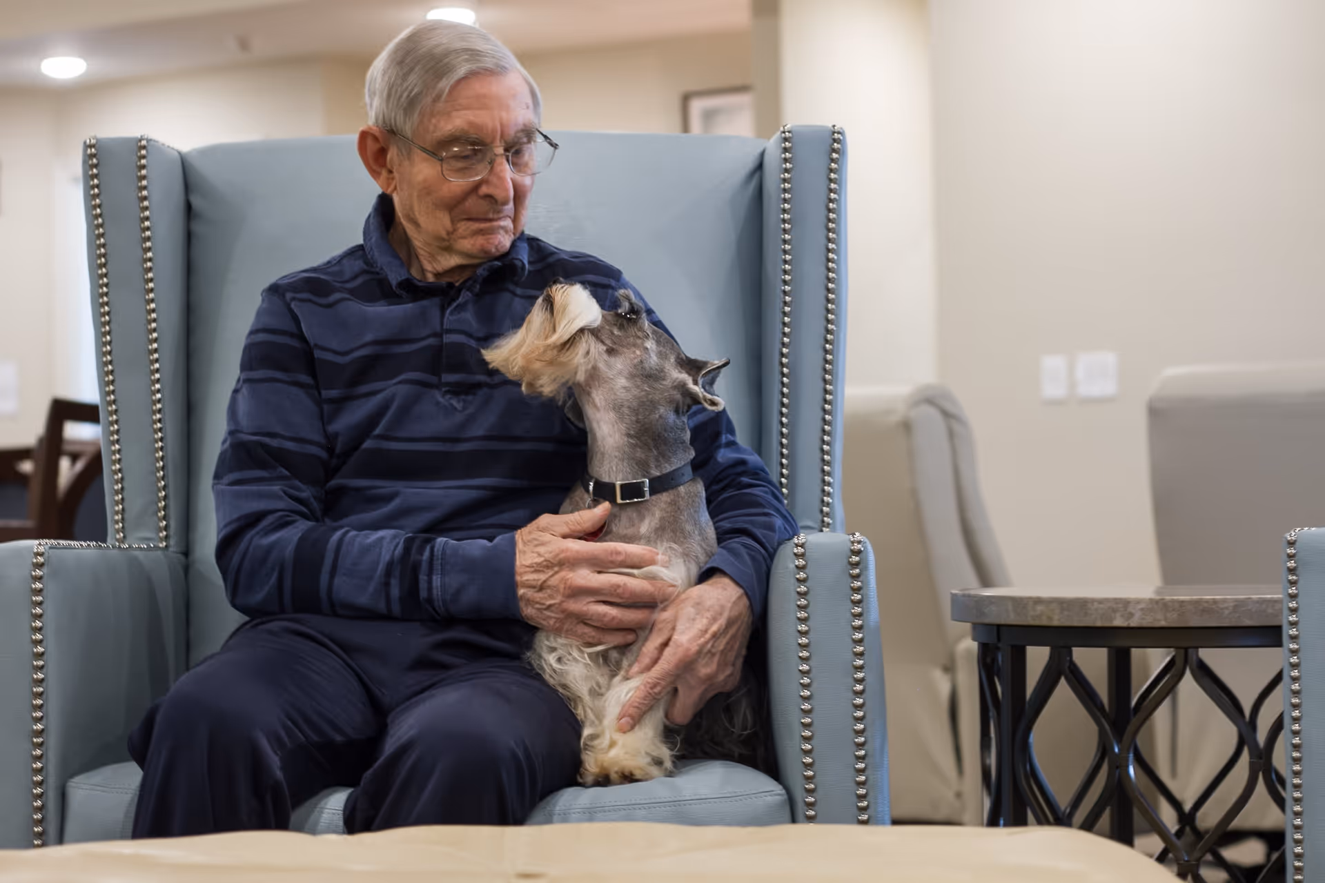 An elderly man wearing glasses and a dark striped shirt sits in a light blue armchair with nailhead trim, holding and looking affectionately at a small dog with a collar. The background shows a neutral-toned room with additional chairs and a side table.