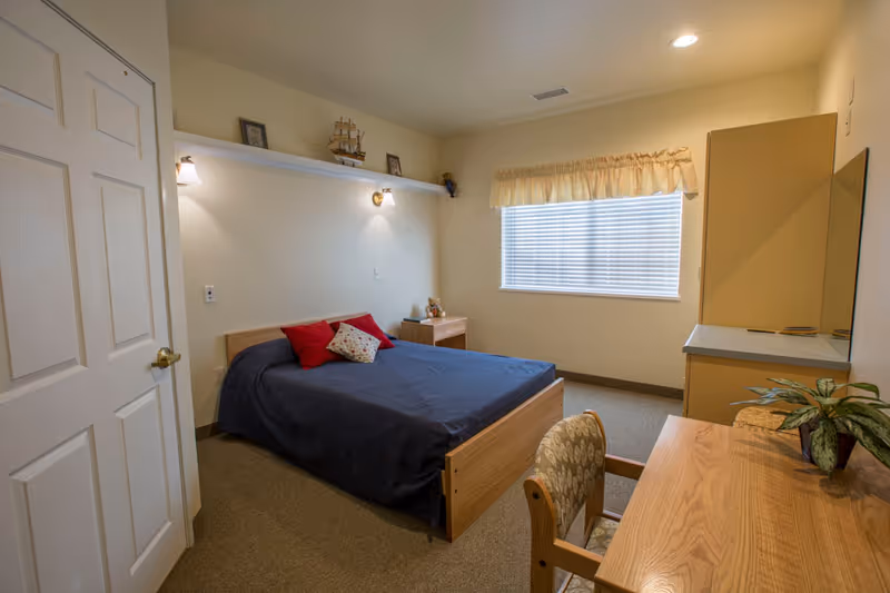 Furnished bedroom with a bed, nightstand, desk and dresser beneath a window with blinds.