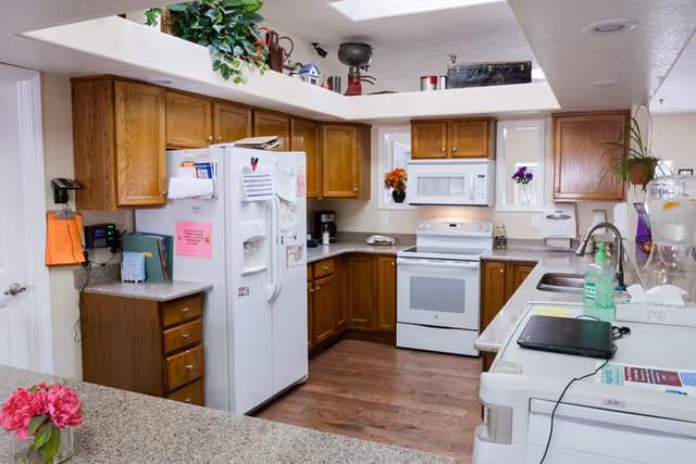 A well-lit kitchen with wooden cabinets, a white refrigerator covered with notes and magnets, a white stove with a microwave above it, and a double sink. The countertops have various items including a laptop, hand sanitizer, a water dispenser, and a vase with flowers. The floor is wooden and there are small windows above the stove with flower pots on the windowsills.