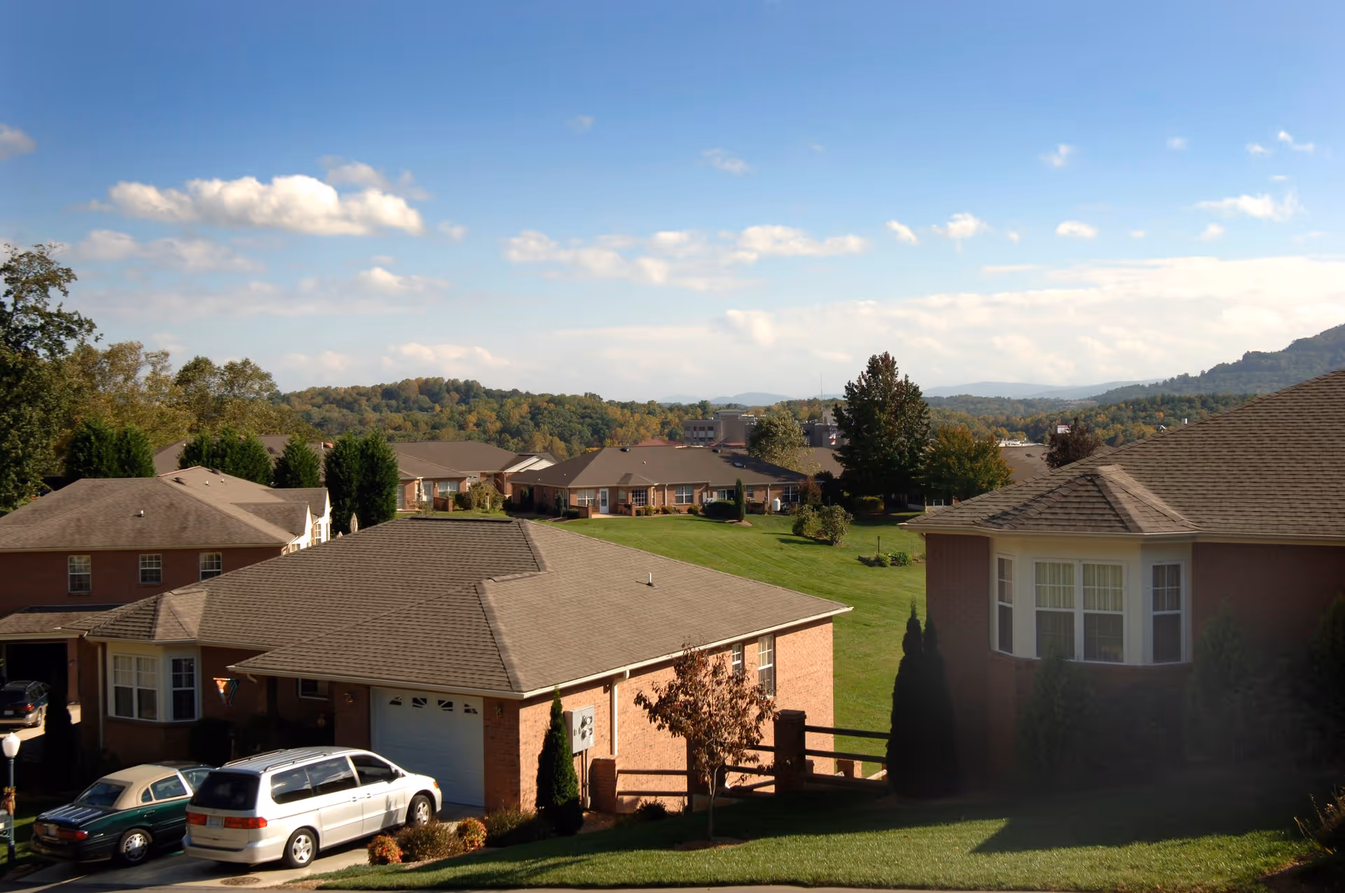 View of a senior living community with brick cottages, lawns, parked cars, and rolling hills under a blue sky.