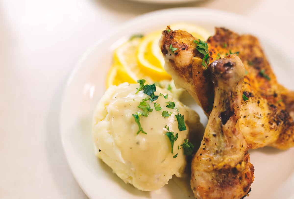 Plate with roasted chicken drumsticks, mashed potatoes topped with gravy and chopped parsley, and lemon slices in the background.