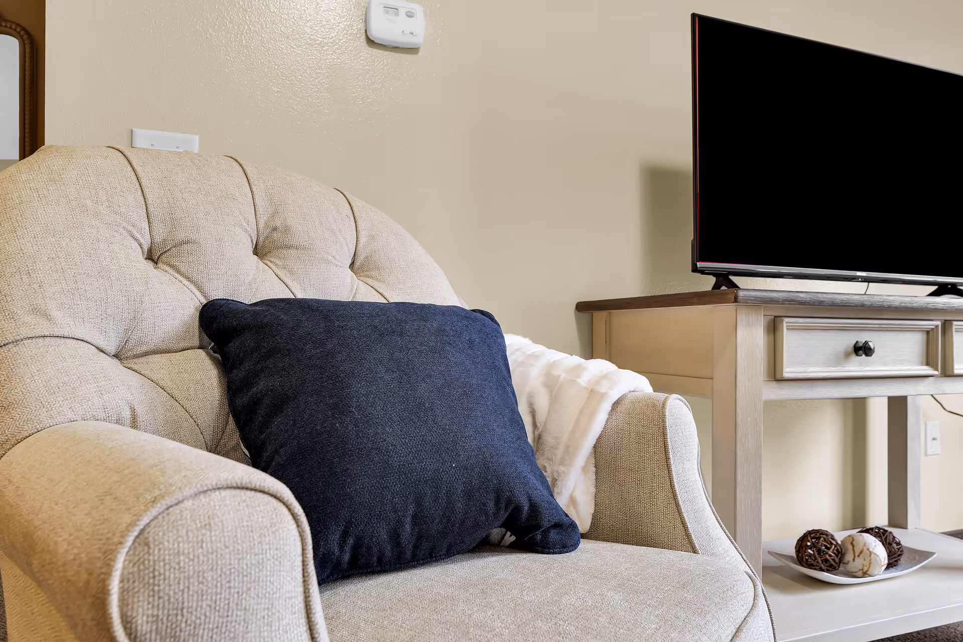 A beige upholstered armchair with a dark blue cushion and a white throw blanket draped over one arm. Next to the chair is a light wooden side table with a flat-screen TV on top and a decorative plate with three decorative balls on the lower shelf. The wall behind is light-colored with a thermostat mounted above the chair.