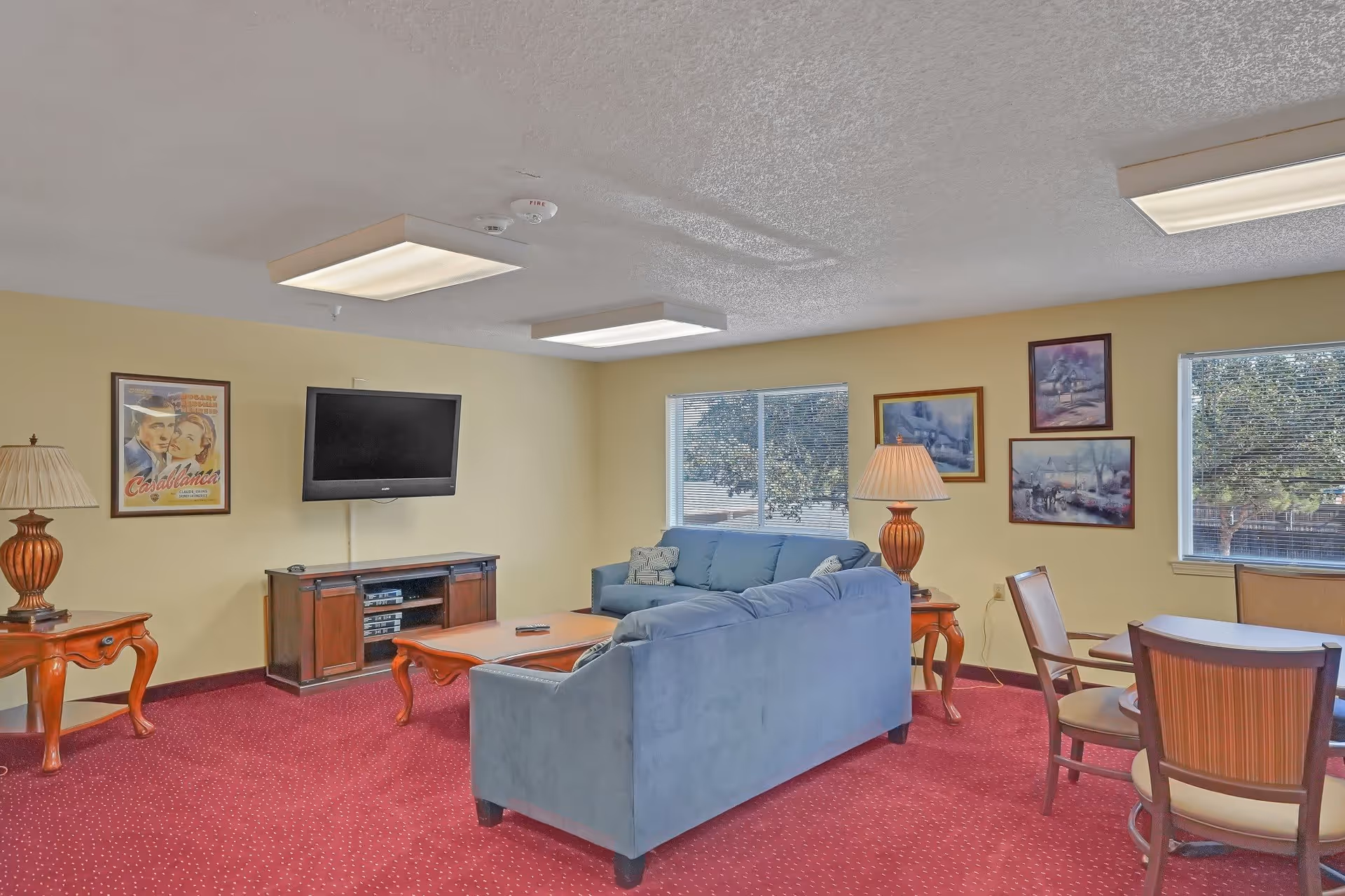 A cozy living room with red carpet, light yellow walls, and two large windows. The room features a blue sofa set, a wooden coffee table, side tables with lamps, a wall-mounted flat-screen TV, and framed artwork on the walls. There is also a small dining table with chairs near one window.