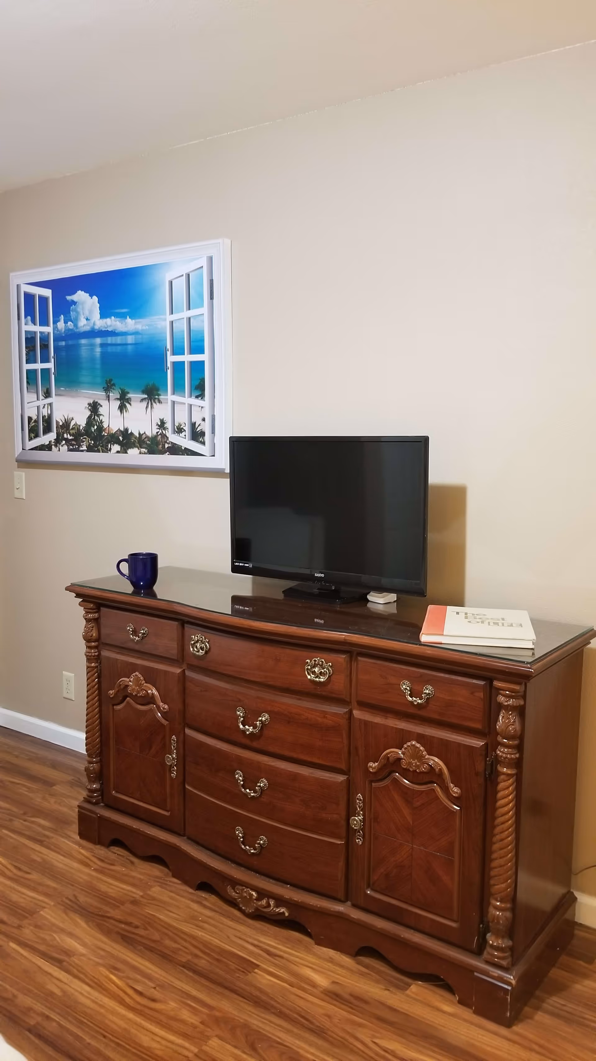 A wooden dresser with ornate handles and carvings, topped with a flat-screen TV, a blue mug, and a book titled 'The Heart of Life'. Above the dresser is a wall decoration resembling an open window showing a tropical beach scene with palm trees and blue sky.