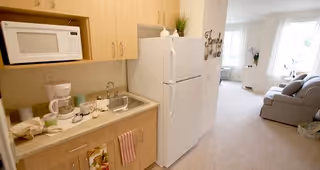 Small kitchenette area with a microwave, coffee maker, sink, and refrigerator. The kitchenette has light wood cabinets and a countertop with cups and a towel hanging. In the background, there is a living room area with a couch, window with white curtains, and some decorative items.