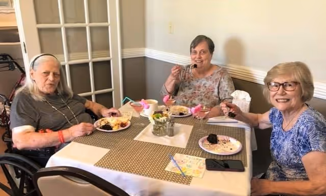Three elderly women sitting around a dining table enjoying dessert. The table is set with plates of cake, drinks in glasses with pink decorations, and a small flower arrangement in the center. The room has light-colored walls with a chair rail and a glass-paneled door in the background.