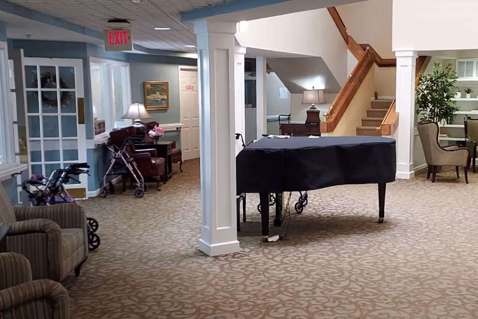 Communal interior lounge with a covered grand piano, seating, a staircase, and several walkers.
