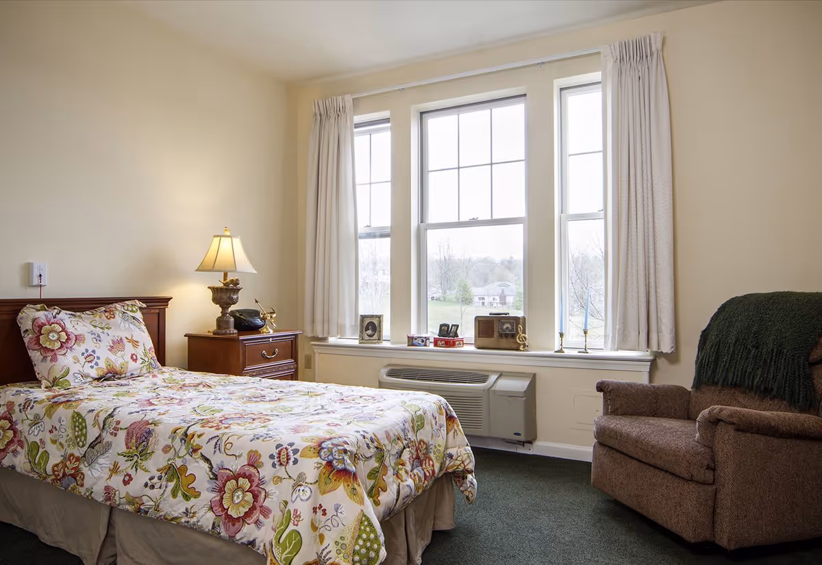 A cozy bedroom with a single bed covered in floral bedding, a wooden nightstand with a lamp and a phone, a large window with white curtains letting in natural light, and a comfortable brown armchair with a green throw blanket.