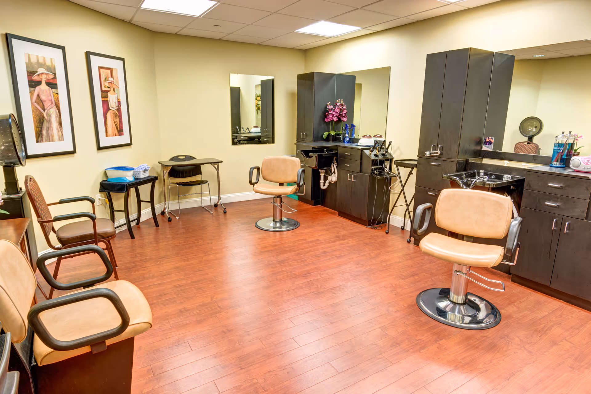 Interior of a salon room with wooden flooring, beige walls, and ceiling lights. The room contains salon chairs, black cabinetry with sinks, mirrors, and decorative framed artwork on the wall.