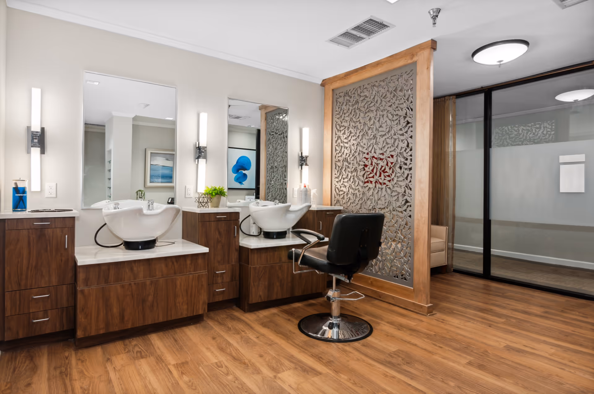 Interior view of a hair salon area in a senior living facility with two white wash basins set into wooden cabinetry, a black salon chair, large mirrors on the wall, vertical light fixtures, and a decorative wooden partition with a leaf pattern. The floor is wood, and there is a glass wall with a door in the background.