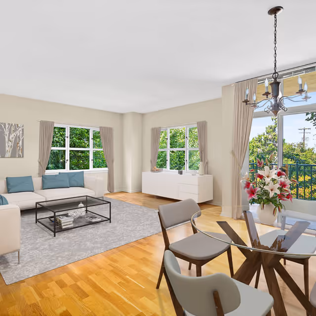 A bright and spacious living room with light-colored walls and wooden flooring. The room features a white sectional sofa with blue cushions, a glass coffee table on a gray area rug, and a white sideboard against the wall. Large windows with beige curtains allow natural light to fill the space, and a glass dining table with four chairs is positioned near a sliding glass door leading to a balcony with greenery outside. A chandelier hangs above the dining table, and a vase with pink and white flowers is placed on the table.